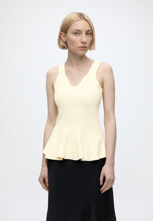 Kvinde med kort blond hår iført en ærmeløs bleg gul peplum top og et sort nederdel mod en ensfarvet lys baggrund.