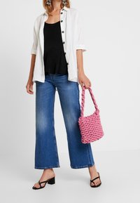 Camisa blanca de botones sobre una blusa peplum negra, combinada con unos pantalones vaqueros de pierna ancha azules y un bolso tejido rosa texturizado. Sandalias negras complementan el look.
