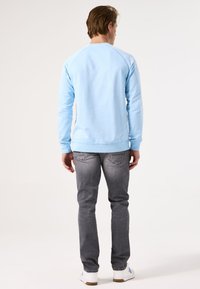 Sudadera azul de manga larga y cuello redondo, combinada con jeans grises y zapatillas blancas, vista desde atrás. Diseño simple y textura suave.