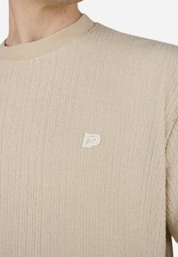 Pull en tricot texturé beige avec un col rond et des finitions côtelées ; présente un petit logo blanc sur la poitrine.