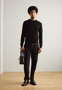 Black long-sleeve shirt, black tapered pants, black leather shoes, holding a small black bag, standing on light wooden floor against a plain wall.