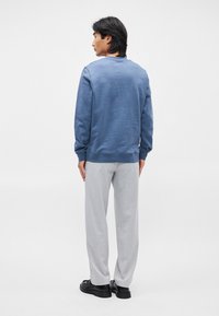 Sweatshirt azul com acabamento texturizado, gola redonda e punhos canelados, combinado com calças cinzentas claras e sapatos pretos. Vista traseira.