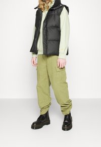 Gilet piumino nero sopra una felpa verde chiaro, abbinato a pantaloni cargo verde oliva e stivaletti neri con lacci. Cuciture e tasche visibili.