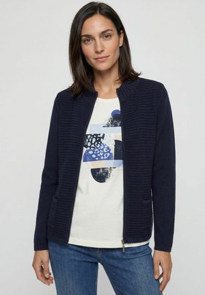 Frau trägt eine marineblaue, gerippte Zip-Up-Jacke über einem weißen T-Shirt mit abstraktem Print und blauen Jeans, steht vor einem einfarbigen hellen Hintergrund.