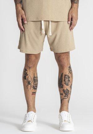 Mand iført beige teksturerede shorts og matchende top, hvide sneakers, med forskellige sorte tatoveringer på begge lægge.