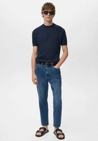 Maglietta a maniche corte a coste color navy, jeans in denim blu, cintura nera e sandali neri. Il modello è in piedi contro uno sfondo bianco.
