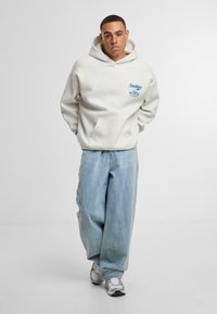 Sudadera con capucha de color gris claro, con un bolsillo canguro en la parte frontal y un logotipo de texto azul, combinada con unos vaqueros anchos de mezclilla azul claro y zapatillas grises.