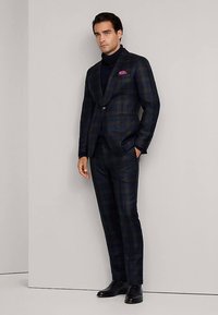 Rutig kostym i mörka toner med gröna och röda accenter, bestående av en kavaj, figurnära byxor och en marinblå polotröja. Inkluderar en näsduk.