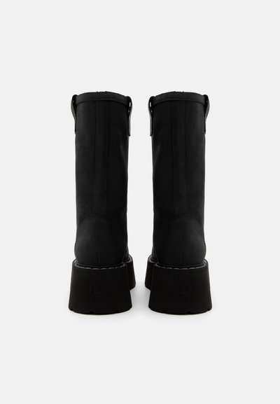 Botas de cuero negro hasta la rodilla con una textura suave y una suela gruesa y acampanada. Presenta tiradores y costuras visibles a lo largo de los bordes.