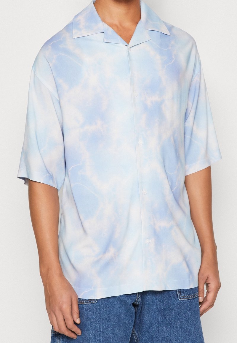 Chemise boutonnée à manches courtes avec un motif de nuages bleu clair et blanc portée avec un jean bleu