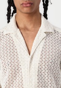 Chemise blanche à motif en zigzag, en tissu léger et texturé, avec un col et une patte de boutonnage.