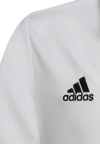 Biała sportowa koszulka z teksturowanej tkaniny. Ma czarną, haftowaną aplikację logo Adidas umieszczoną po lewej stronie klatki piersiowej.