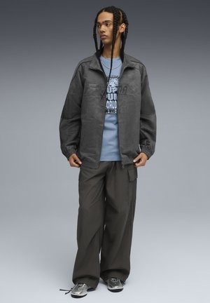 Veste zippée grise avec une texture douce, assortie à un t-shirt graphique bleu clair et un pantalon large sombre ; modèle portant des chaussures métalliques.