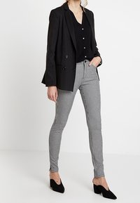 Černý dvouřadý blazer přes černý top, spárovaný s černobílými kostkovanými slim-fit kalhotami a černými podpatky.