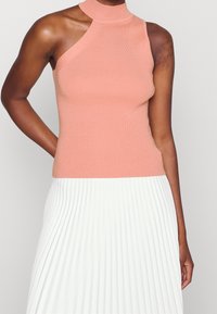 Top sin mangas de canalé en rosa claro con un cuello alto y un corte asimétrico en el hombro, combinado con una falda blanca con pliegues.