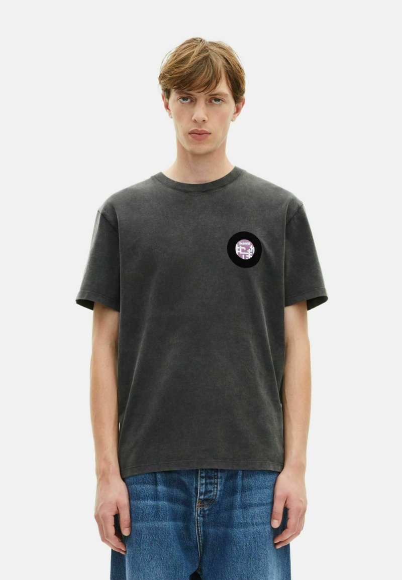 Temno siva bombažna majica s kratkimi rokavi in krožnim izrezom, ki se ponaša s krožno vijolično oznako na levi prsni strani.