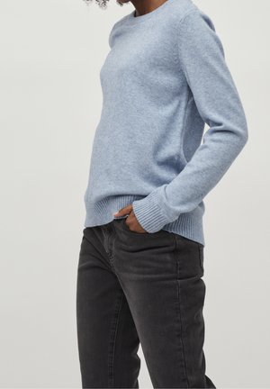 Persona con un suéter de manga larga de color azul claro y pantalones gris oscuro con una mano en el bolsillo, mostrada desde los hombros hasta los muslos.