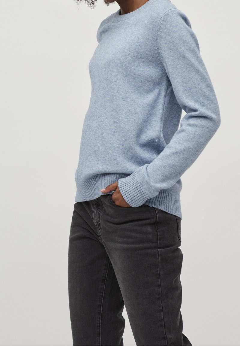 Personne portant un pull bleu clair à manches longues et un pantalon gris foncé, une main dans la poche, montrée des épaules aux cuisses.