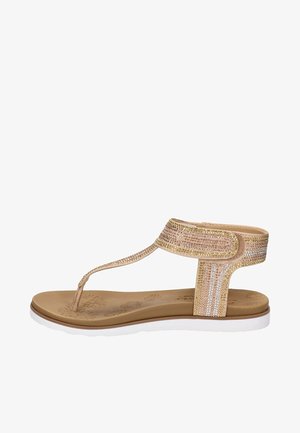 Sandalia plana beige con tira alrededor del tobillo y entre los dedos, adornada con pequeñas piedras brillantes y suela de goma blanca.