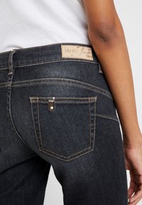 Dark denim jeans with a fitted design. Features a branded leather patch, contrast stitching, and a small zippered pocket on the back.