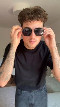 Ochelari de soare negri cu lentile rotunde ținuți în mâini, cu accente argintii, purtați împreună cu un tricou negru cu mâneci scurte și însoțiți de tatuaje pe brațe.