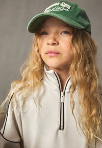Green cap with embroidered text, light gray zip-up sweatshirt with black trim, and wavy, long hair. Neutral background.