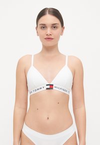 Bralette bianco a forma di triangolo con spalline sottili e regolabili, fascia con logo che presenta accenti rossi, bianchi e blu, e una texture liscia e lucida.