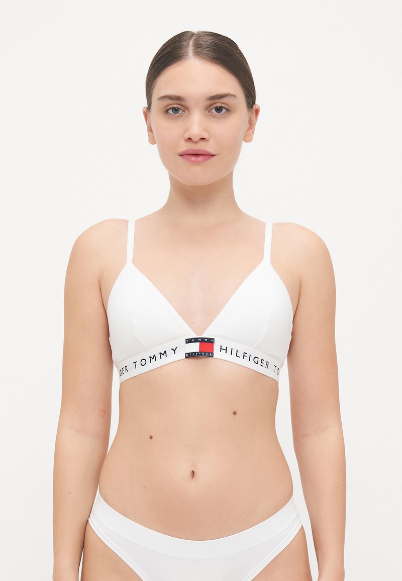 Bralette bianco a forma di triangolo con spalline sottili e regolabili, fascia con logo che presenta accenti rossi, bianchi e blu, e una texture liscia e lucida.