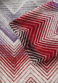 Alfombra texturizada con patrones en zigzag en tonos rojos, blancos y morados, que muestra colores contrastantes y una superficie tejida y suave.