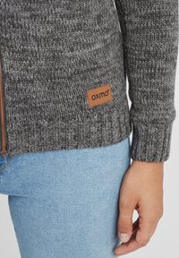 Gray knitted cardigan with ribbed cuffs and hem, featuring a brown leather label with "oxmo." Paired with light blue denim jeans.