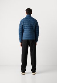 Blauwe pufferjack met een doorgestikt ontwerp, hoge kraag en elastische manchetten. Draagt met zwarte broek en sneakers. Gladde textuur en lichtgewicht uitstraling.