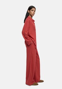 Kobieta stojąca bokiem, ubrana w luźną, czerwoną bluzkę z długim rękawem i szerokie spodnie, w brązowych butach, na białym tle.