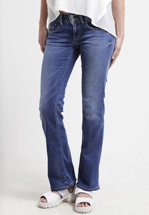 Femme portant un jean bleu taille mi-haute avec fermeture à double bouton et des sandales blanches à plateforme, debout sur un fond blanc.