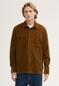 Brown corduroy shirt with a collared neck, button closure, and two chest pockets. Textured fabric with vertical ridges. Long sleeves.