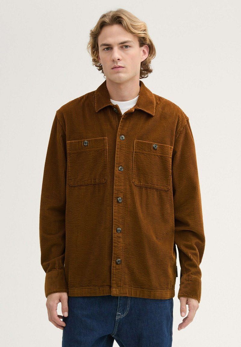 Brown corduroy shirt with a collared neck, button closure, and two chest pockets. Textured fabric with vertical ridges. Long sleeves.