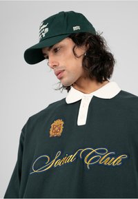 Polo de color verde oscuro con un cuello blanco, que presenta texto bordado en oro y azul, así como un escudo en el pecho. Combinado con una gorra a juego.