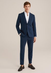 Mlad čovjek stoji odjeven u tamnoplavi odijelo, bijelu košulju i tamnosmeđe cipele na loafere, naslonjen na jednostavnu bež pozadinu.