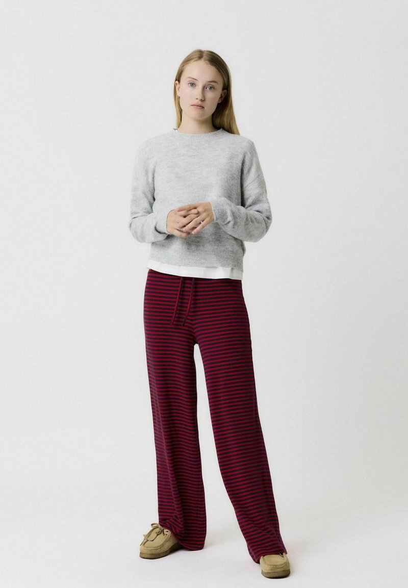 Un pull gris à col rond, associé à un pantalon large rayé rouge. La tenue présente une coupe décontractée et des textures douces. Des chaussures brunes à enfiler.