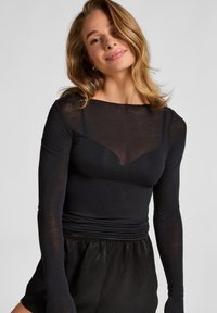 Jonge vrouw met blond haar, gekleed in een doorzichtig zwart langemouwen T-shirt over een zwart hemd en zwarte high-waist onderkleding, die zachtjes glimlacht.