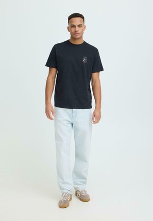 Nuori mies seisoo päällään musta graafinen t-paita, vaaleansiniset farkut ja beiget lenkkarit yksinkertaista vaaleaa taustaa vasten.