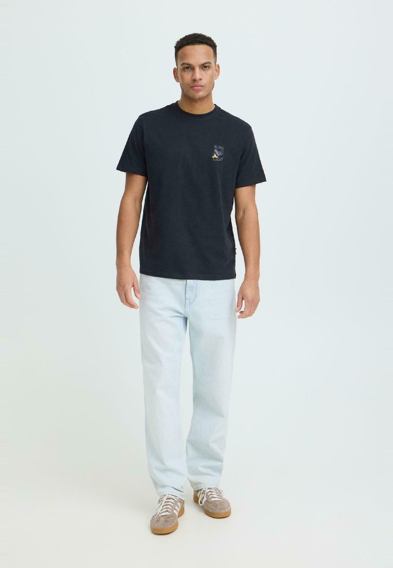 Νέος άνδρας που στέκεται, φορώντας μαύρο μπλουζάκι με σχέδιο, ανοιχτόχρωμο τζιν και μπεζ αθλητικά παπούτσια, μπροστά σε απλό φωτεινό φόντο.