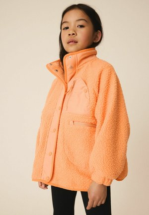 Junges Mädchen trägt eine hellorange Fleecejacke mit Druckknöpfen und Reißverschlusstaschen, steht vor einem schlichten beigen Hintergrund.