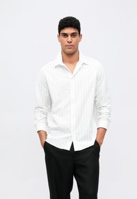 Hvit langermet skjorte med tynne vertikale striper, knappet ned, kombinert med mørke skreddersydde bukser. Glatt stoff, klassisk kragedesign.