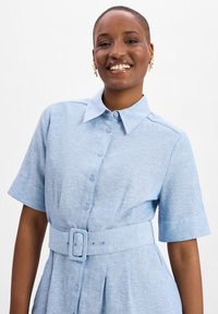 Light blue short-sleeve shirt dress made of textured fabric, featuring a collared neck, button front, and a belted waist with a rectangular buckle.