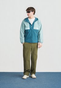 Νεαρός άνδρας με σκούρα γυαλιά ηλίου, τιρκουάζ μπουφάν με κουκούλα, χακί παντελόνια cargo και λευκά αθλητικά παπούτσια, στέκεται σε μπλε χαλί μπροστά σε λευκό τοίχο.