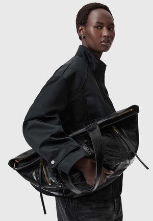 Moteris juoda striuke laiko didelę juodą odinę rankinę su auksinėmis užtrauktukais, fone vientisa pilka spalva.