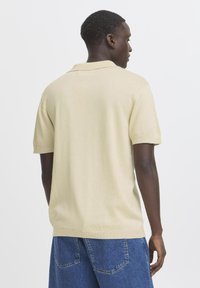 Beige rövid ujjú pólóing klasszikus gallérral, kötött textúrával és bordás mandzsettával, kék farmer rövidnadrággal viselve.