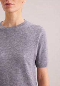 Hellgrauer kurzärmeliger Strickpullover mit Rundhalsausschnitt, weicher Textur, dezenten Sprenkeln sowie gerippten Bündchen und Saum. Nahaufnahme, minimalistischer Hintergrund.