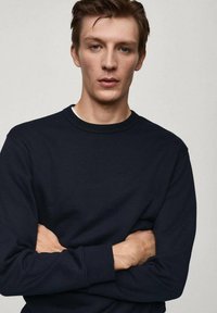 Felpa girocollo blu navy con maniche lunghe. Tessuto liscio, polsini e bordo a coste. Modello con le braccia incrociate che si trova di fronte a uno sfondo neutro.