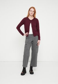 Cardigan bordeaux con chiusura a bottoni, indossato sopra una maglietta bianca. Pantaloni cargo grigi con tasche e stivali neri con lacci completano l'outfit.
